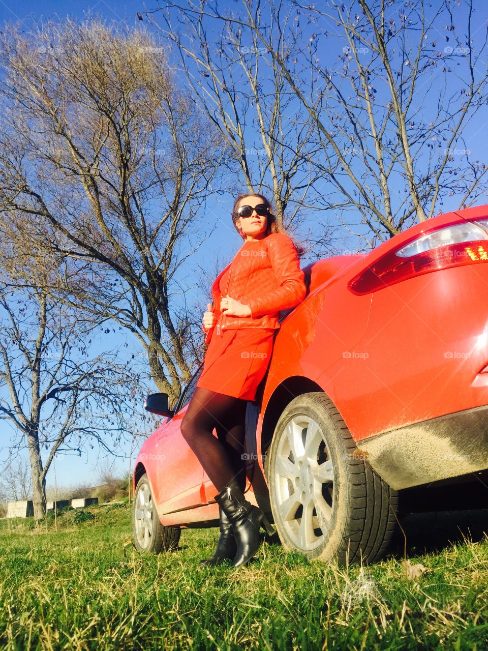 Woman in red dress standing near red car with blue sky and green grass in the background 