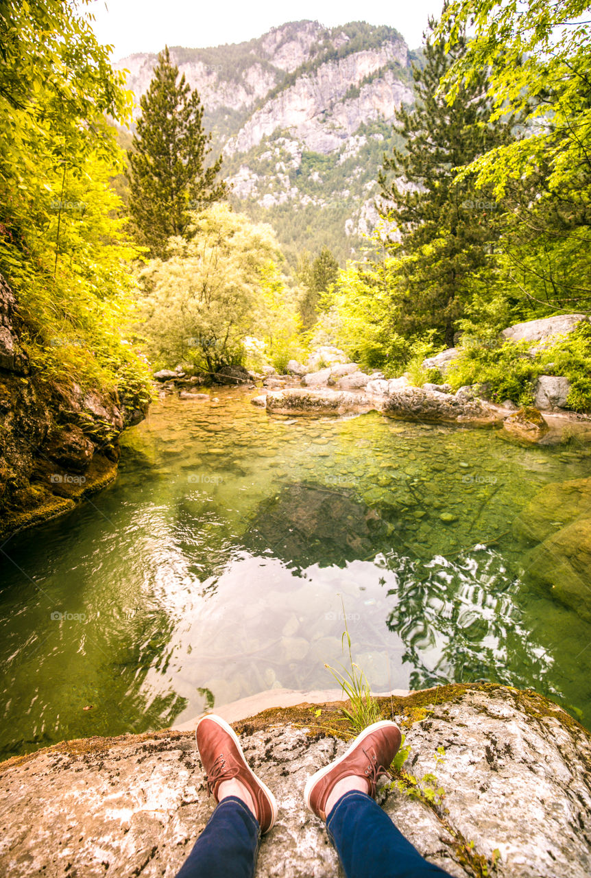 Feet in brown shoes sitting on the rock by the river in front of a gorgeous mountain landscape at Olympus mountain in Greece.