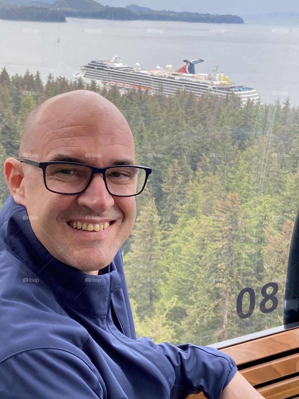 A bald man with glasses sitting by a window with a view of a Carnival Cruise ship in Alaska.