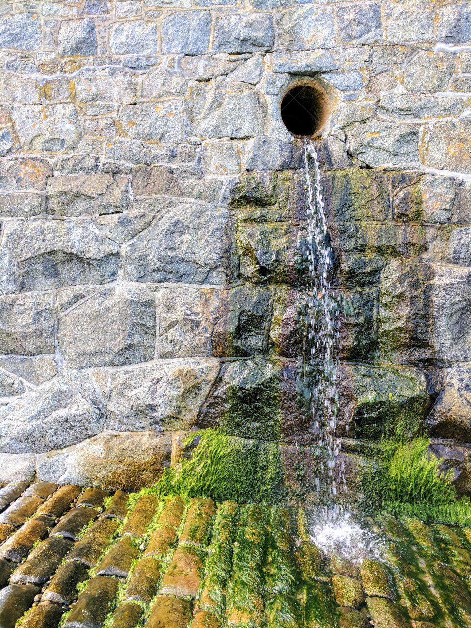 Water pouring out from an outlet pipe and cascading down the side of a granite stone wall.