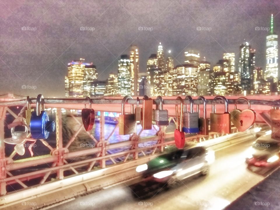 urban skyline at night from Brooklyn bridge. foreground features love locks hanging from bridge with nonstop traffic below