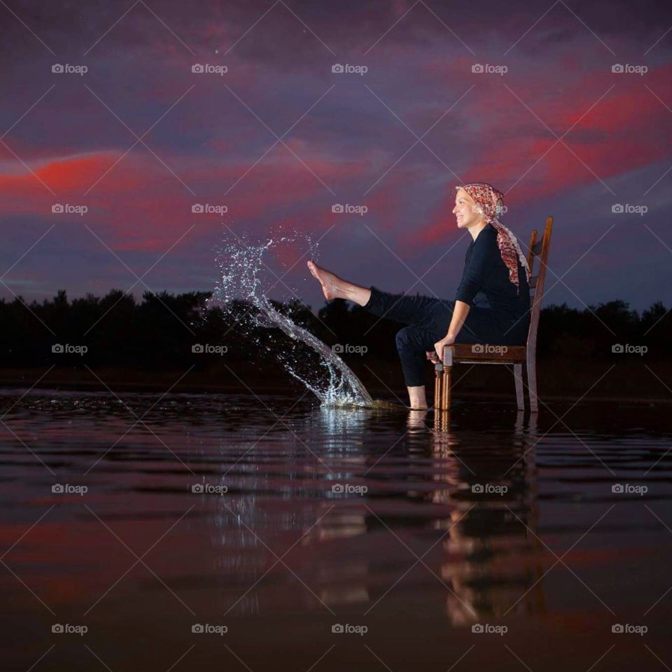 Beautiful young blonde woman sitting on a chair in the water of a lake around Dusk,sunset, playfully kicking around the water with her feet