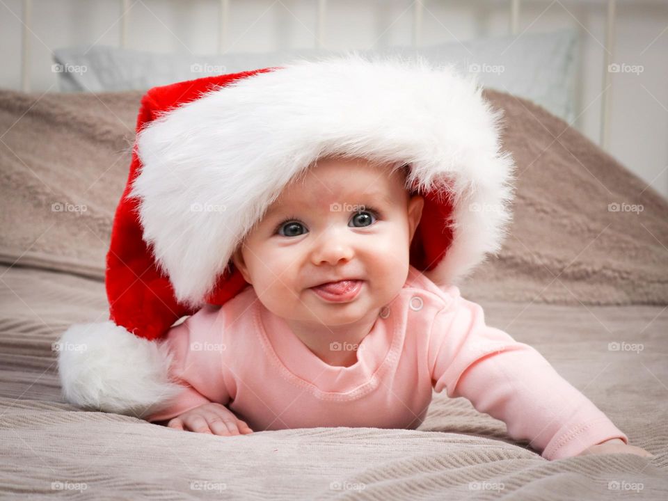 Portrait of a beautiful little happy caucasian baby girl in a santa claus hat lying on the bed with tongue hanging out,close-up side view. Children emotion concept, happy childhood.