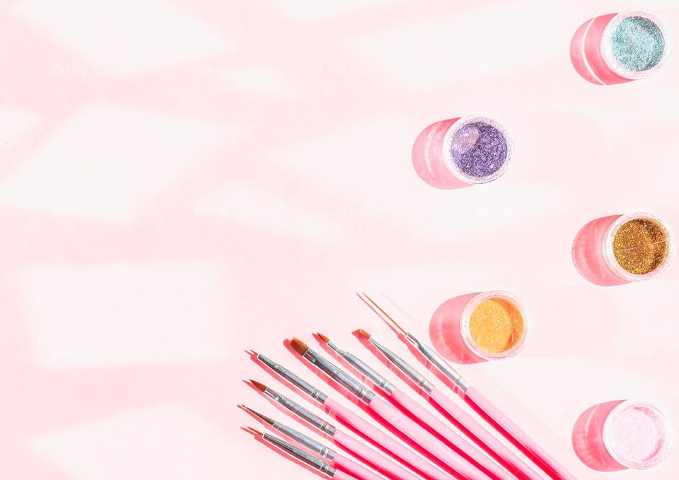 A set of nail brushes and multicolored glitter in mini jars lie on the right on a pink background with glare from the sun and a copy of the space on the left, flat lay. The concept of cosmetics and beauty