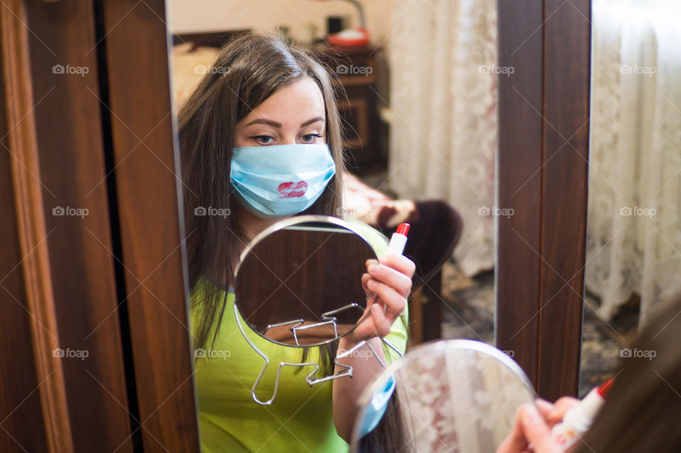 A young girl does makeup for a walk outside in a protective mask against the coronavirus pandemic, her lips are painted with red lipstick.