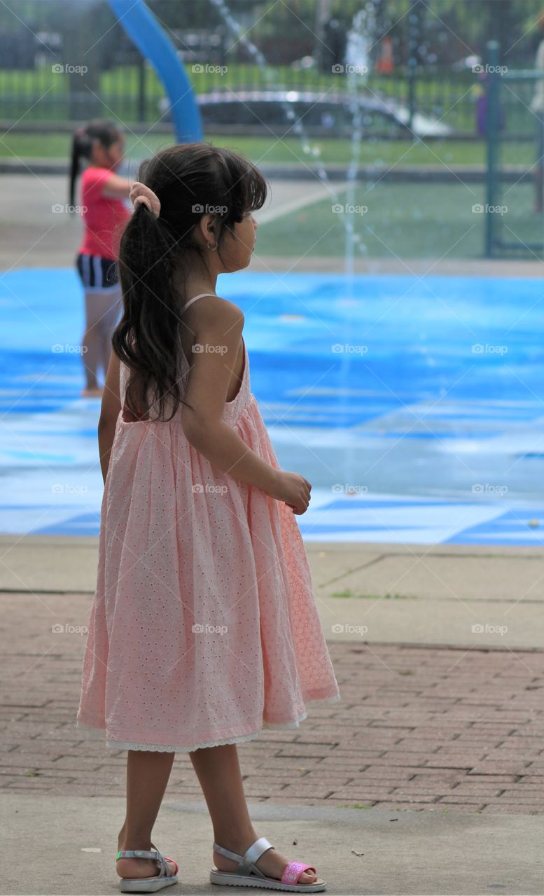 Little girl in long light pink dress and sandals with long hair tied back in waterpark looking at water sprays 