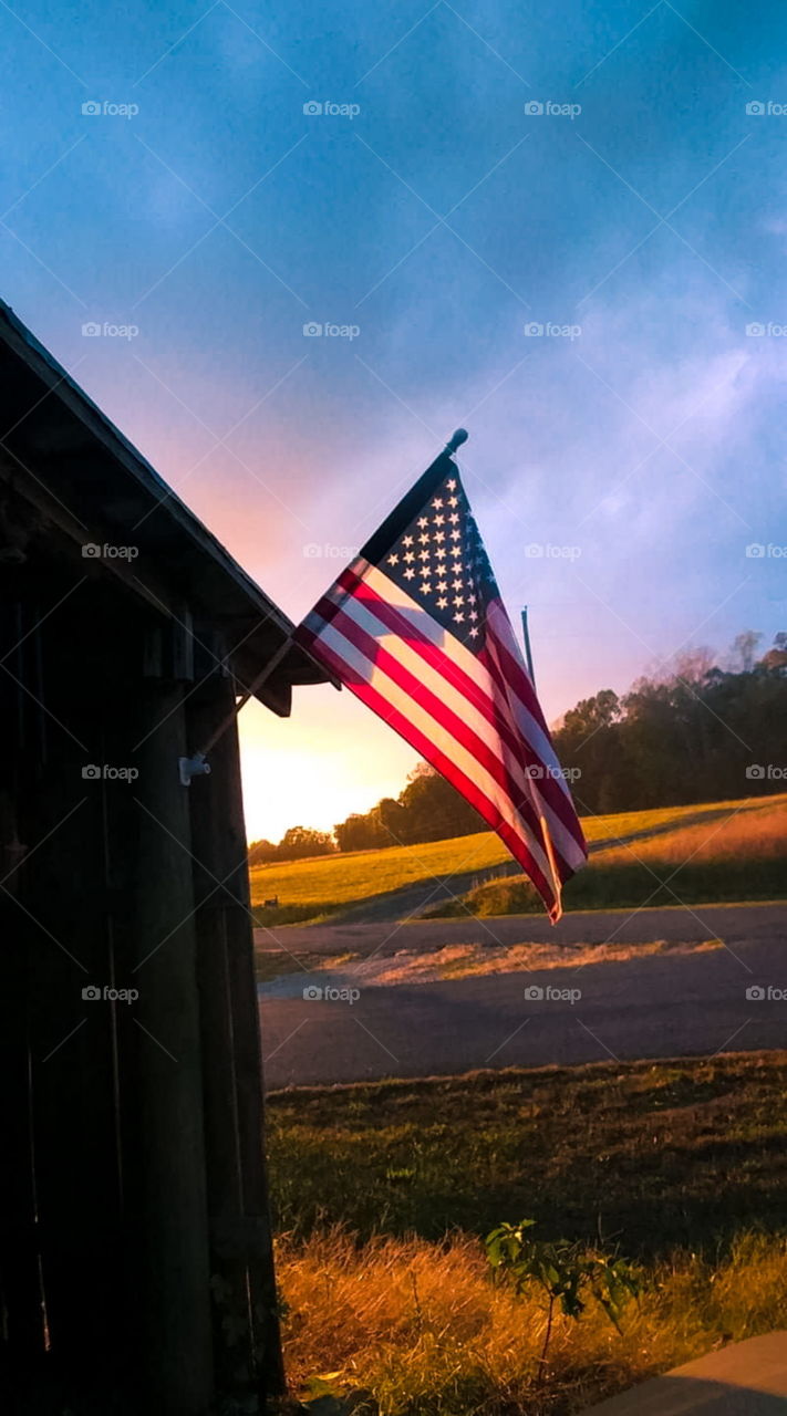 American Flag hanging on side of old tobacco barn with Sun set illuminating flag. Overlooking small country road with hay fields lit in the sun's glow.