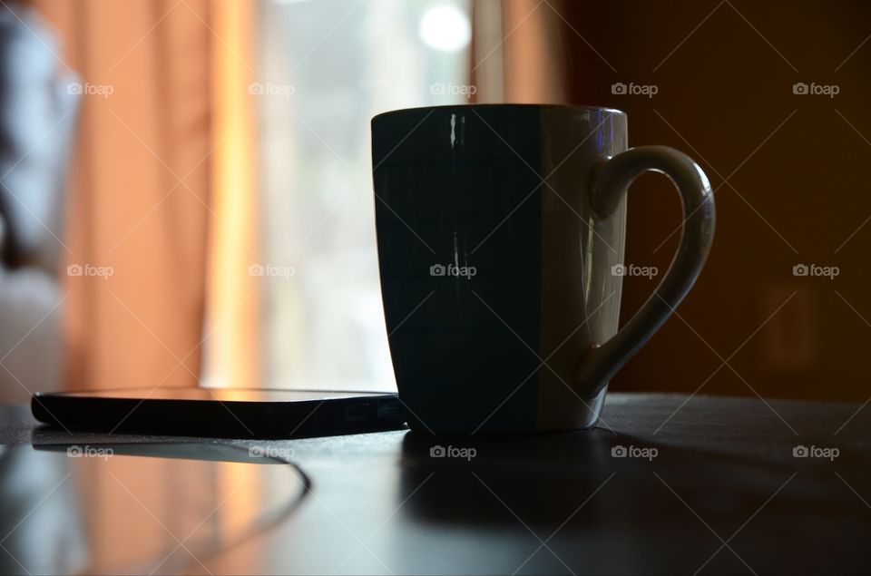 A hot cup of coffee and a cellphone rest on a livingroom table in the morning sunday light.