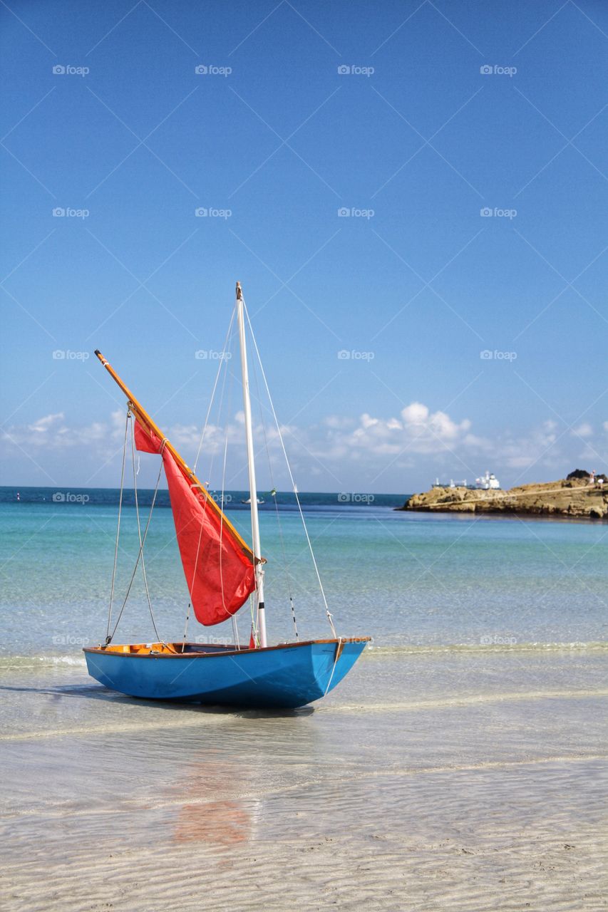 Little Red Sail Boat. A small blue boat with a bright red sail on a sandy beach during a lovely summer day.