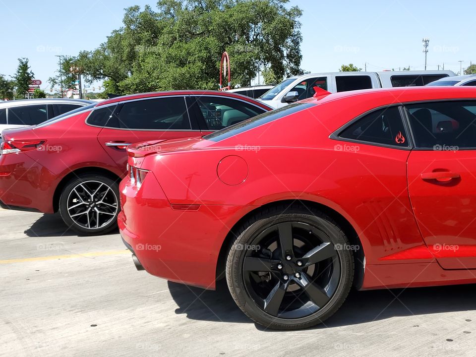 Two red sports cars parked next to each other in a parking lot.  Including a red Camaro