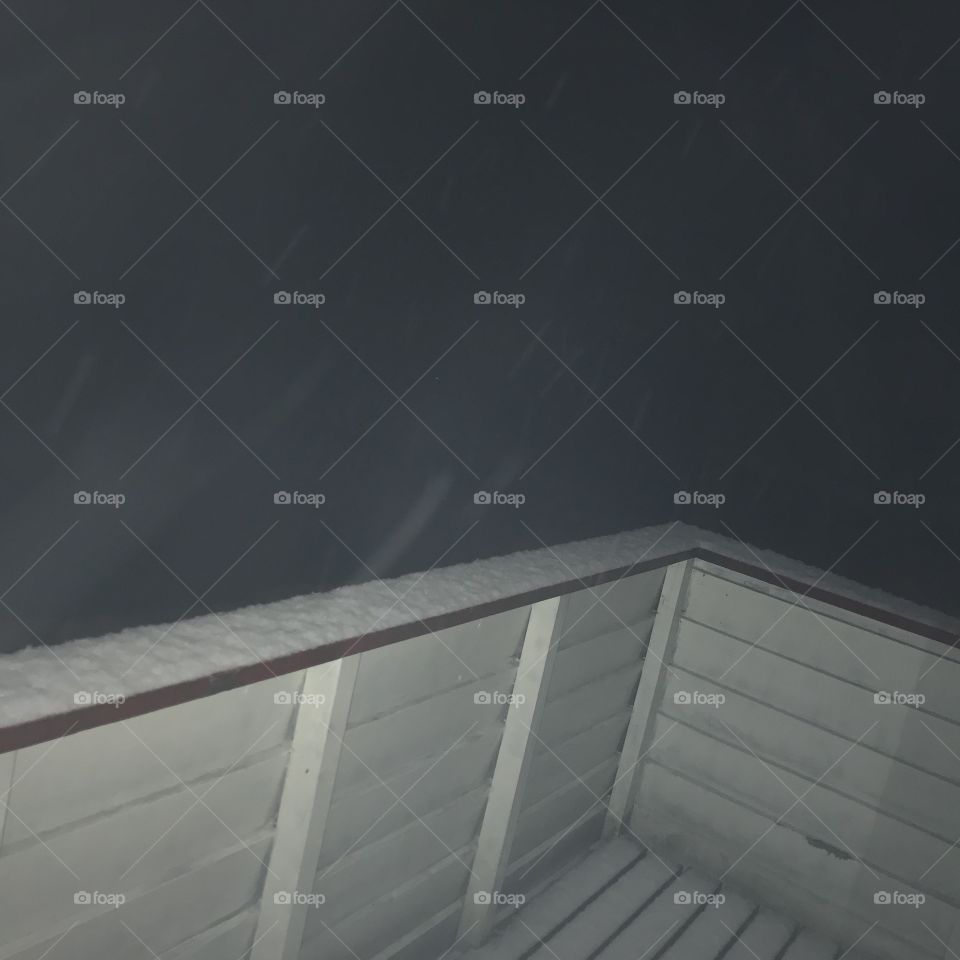 You can only see the fence, and the white snow laying on top of it. It is too dark to see any further out in the evening darkness. The fence is colored white and red, but most of the red is covered in snow. This is taken at wintertime in Norway.