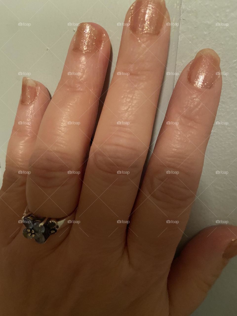 Natural looking nails in pinkish gold sparkly polish against a white wall.