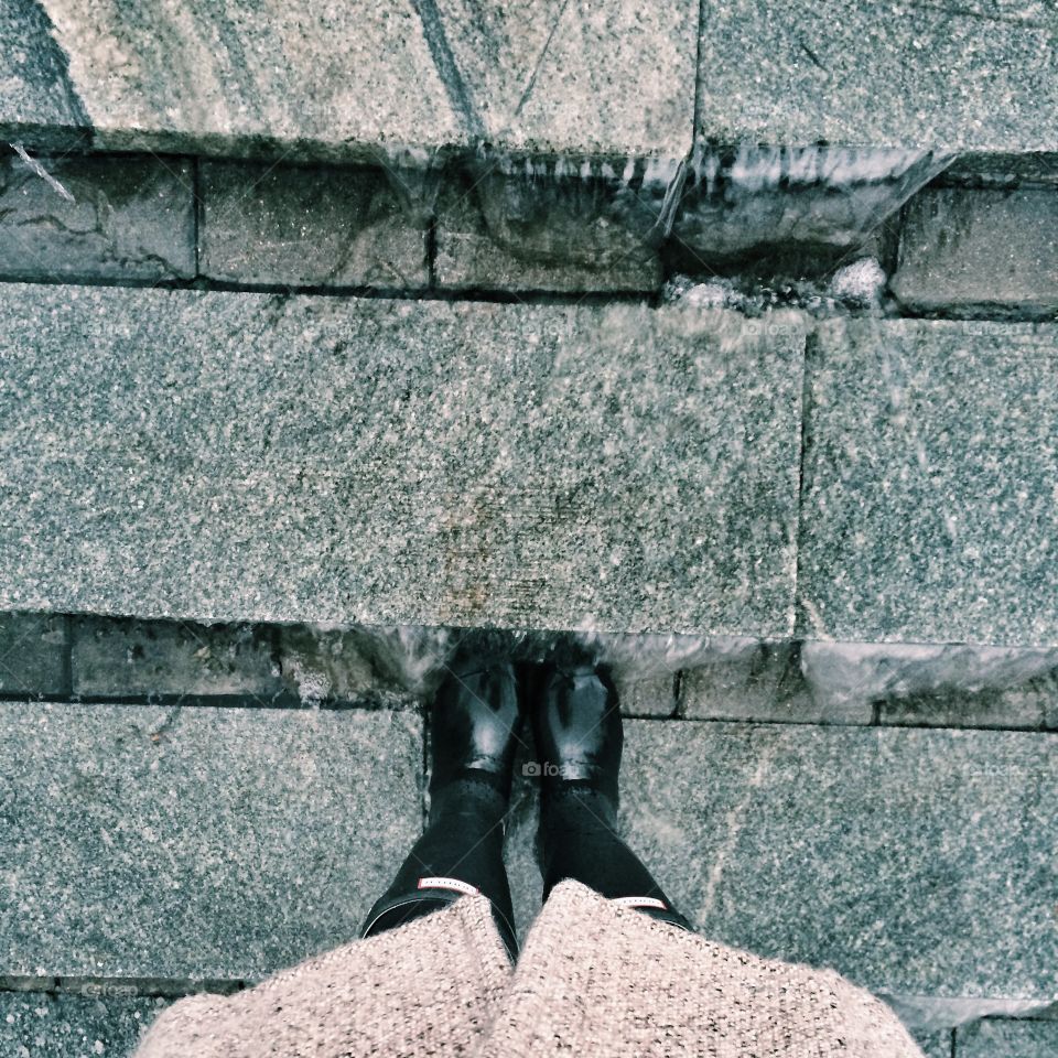 Girl in black rubber hunter boots and a grey wool coat standing with her legs on marble stairs in a rainy autumn weather when water is running 