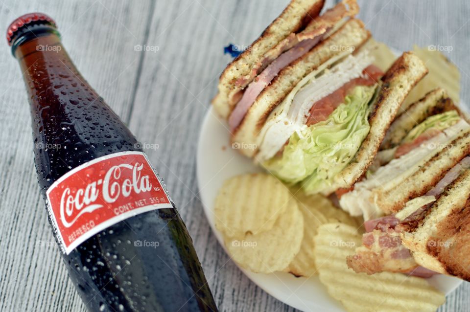 A can and a Bottle of Coca Cola with a club sandwich on a white rustic background 