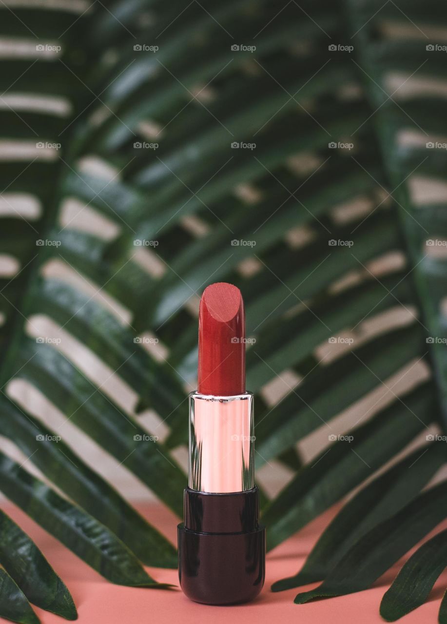 One red lipstick stands on a rose background with palm branches on the back of a white brick wall, close-up side view. The concept of female cosmetics, beauty salon.