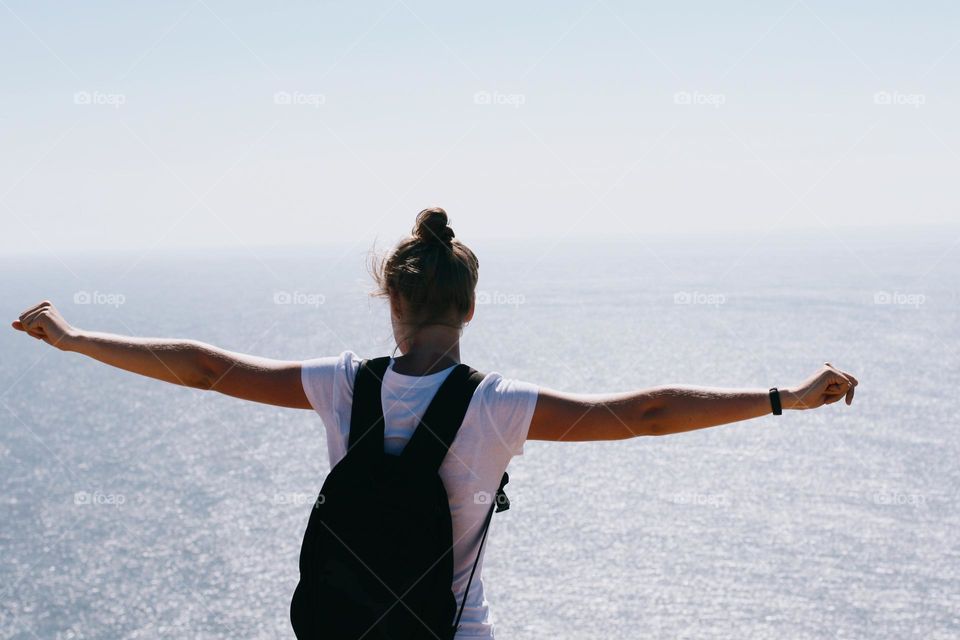 Young girl with a backpack table on top of a mountain with her arms outstretched against the ocean on a sunny summer day, back view 