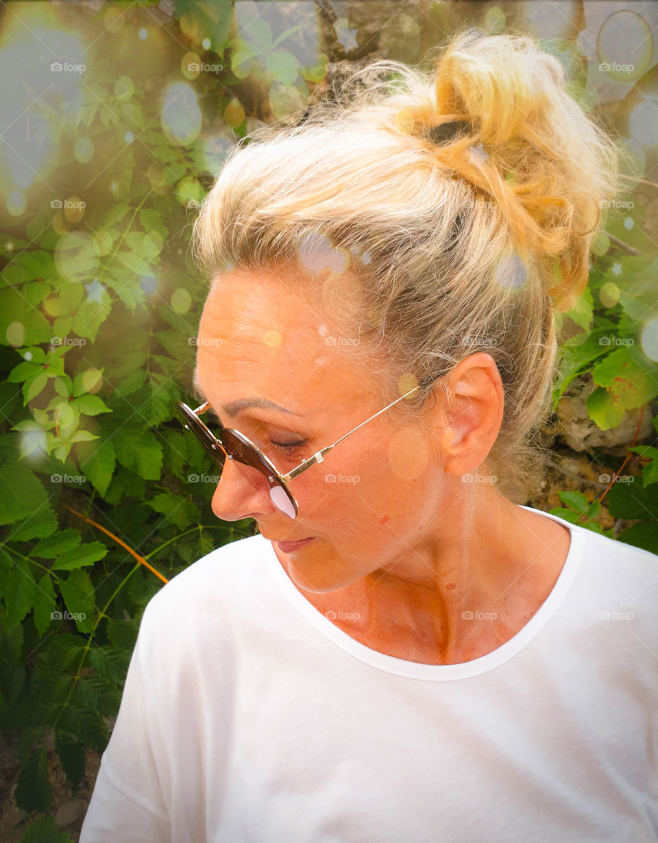 Portrait of a smiling woman in profile in round sunglasses and a white T-shirt on a green garden.  Lovely aged woman