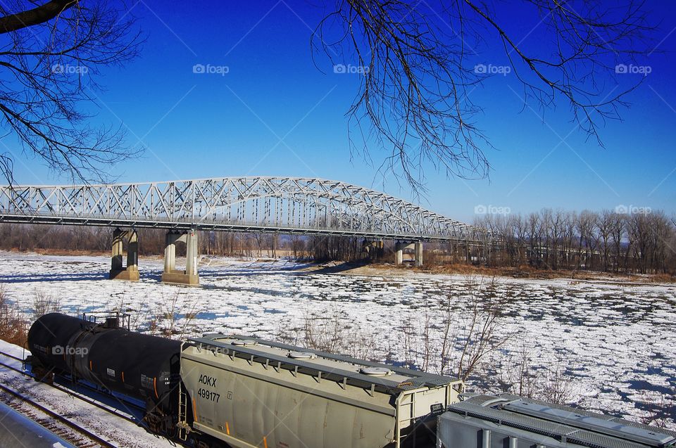 Logistics and transportation image with frozen Missouri River, road bridge and train on a clear winter day with snow and ice on ground 