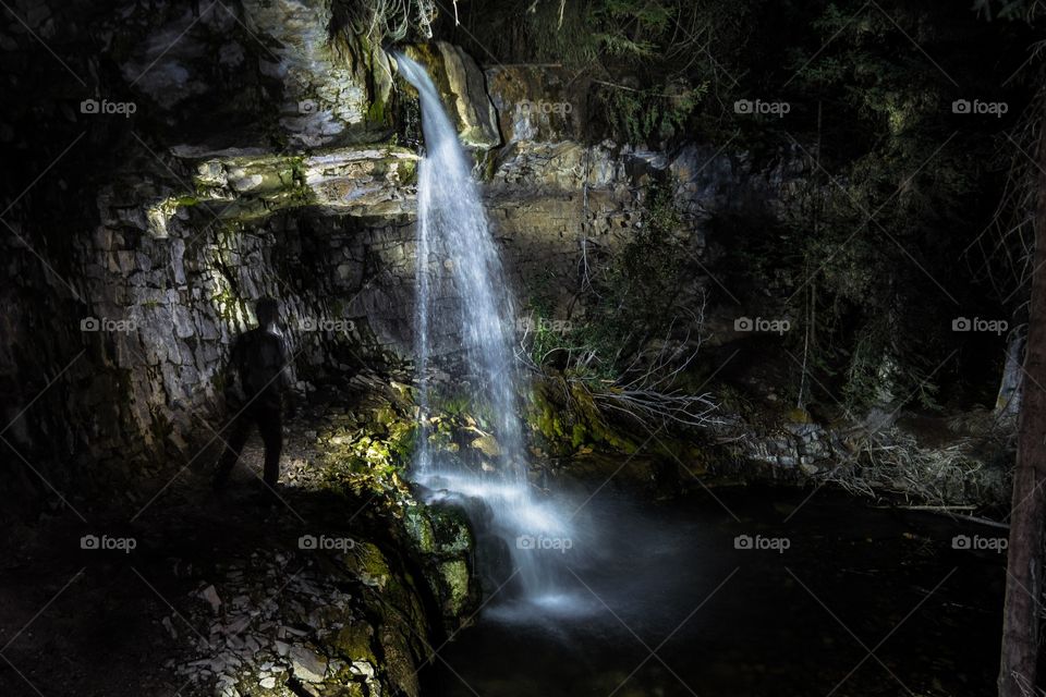 Standing beside an overhanging waterfall at night, in a canyon with cliffs, a pond, and a path leading behind the falls, in Kananaskis, Alberta, Canada 