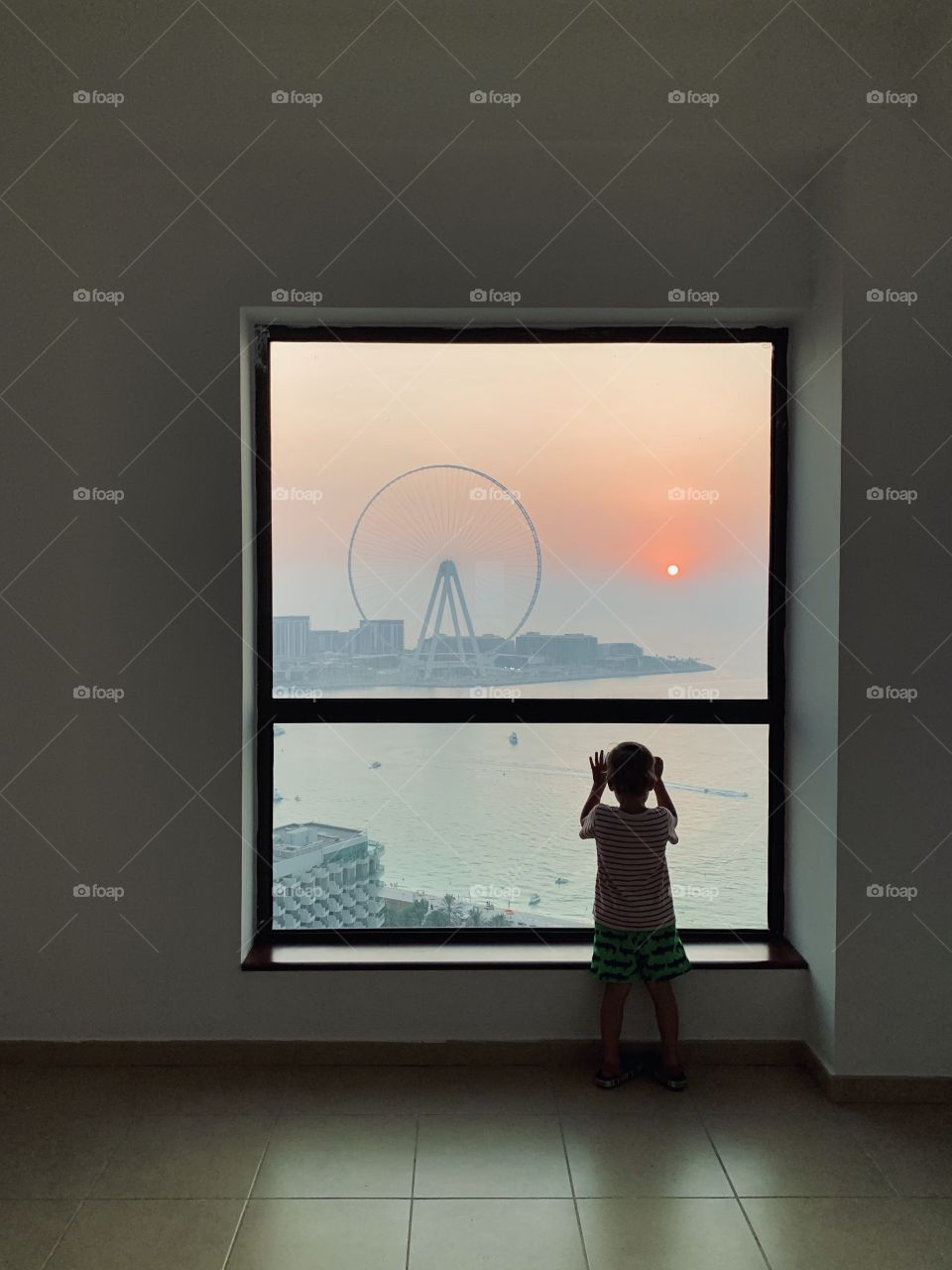 Little boy near window with scenic view of Ferris wheel at sunset 