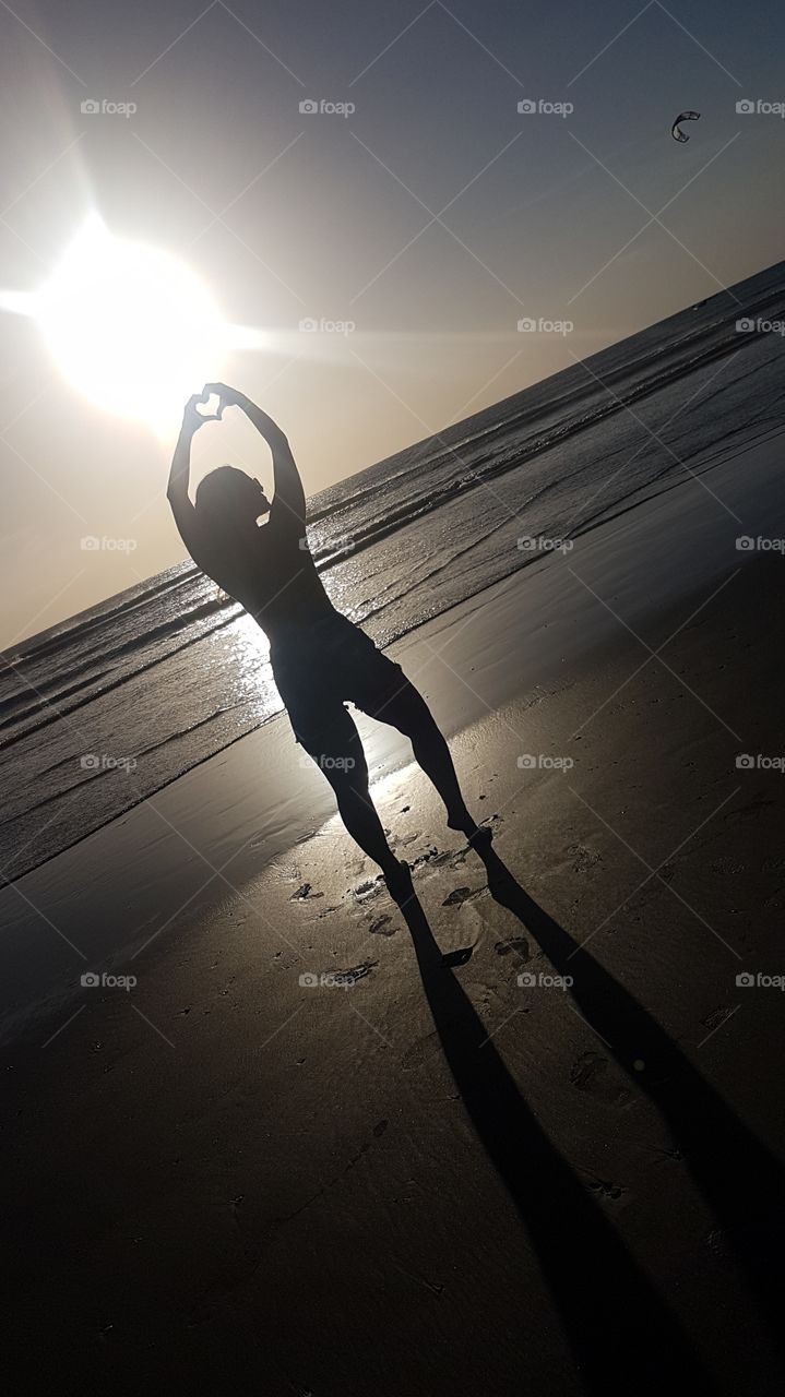 silhueta e coração contra o sol, em frente ao mar.