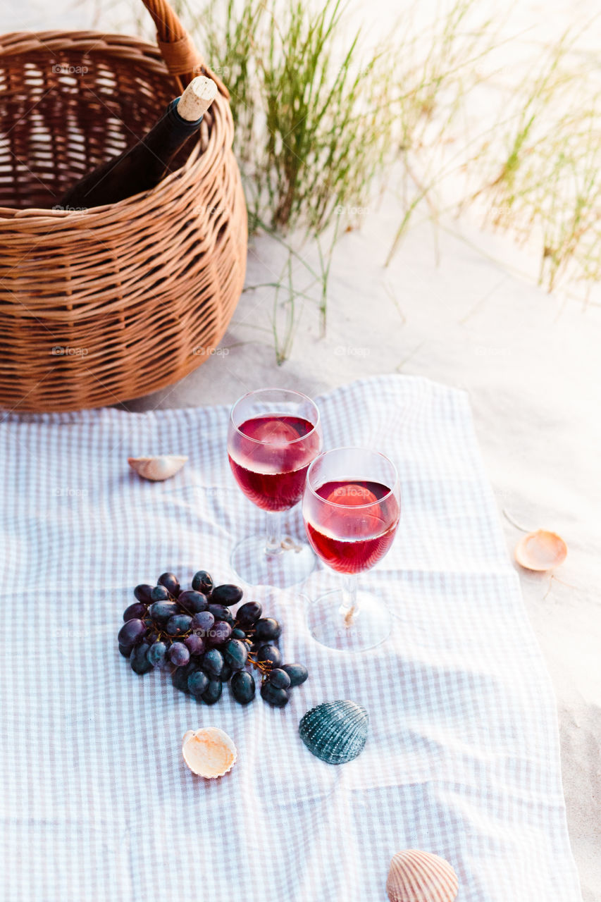 Two wine glasses with red wine standing on cloth, on beach, beside grapes and wicker basket with bottle of wine