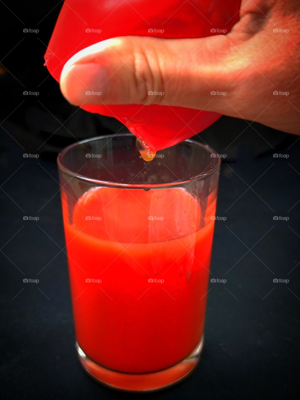 Glassful with red tomato juice. Above the glass is a female hand, which squeezes the tomato. Drop of tomato juice. Black background