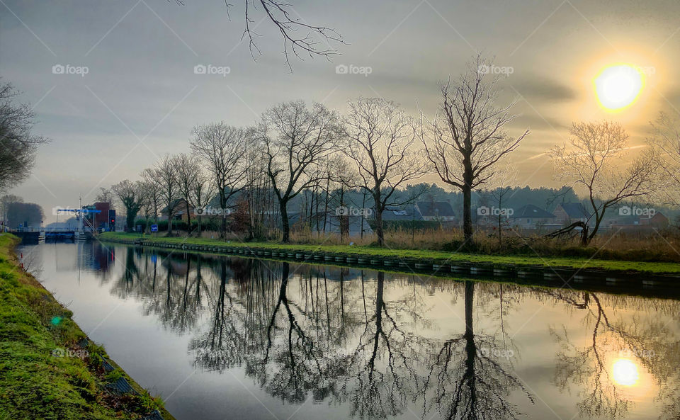 Winter landscape of bare trees under a grey cloudy sunrise sky reflected in the water of a canal or river