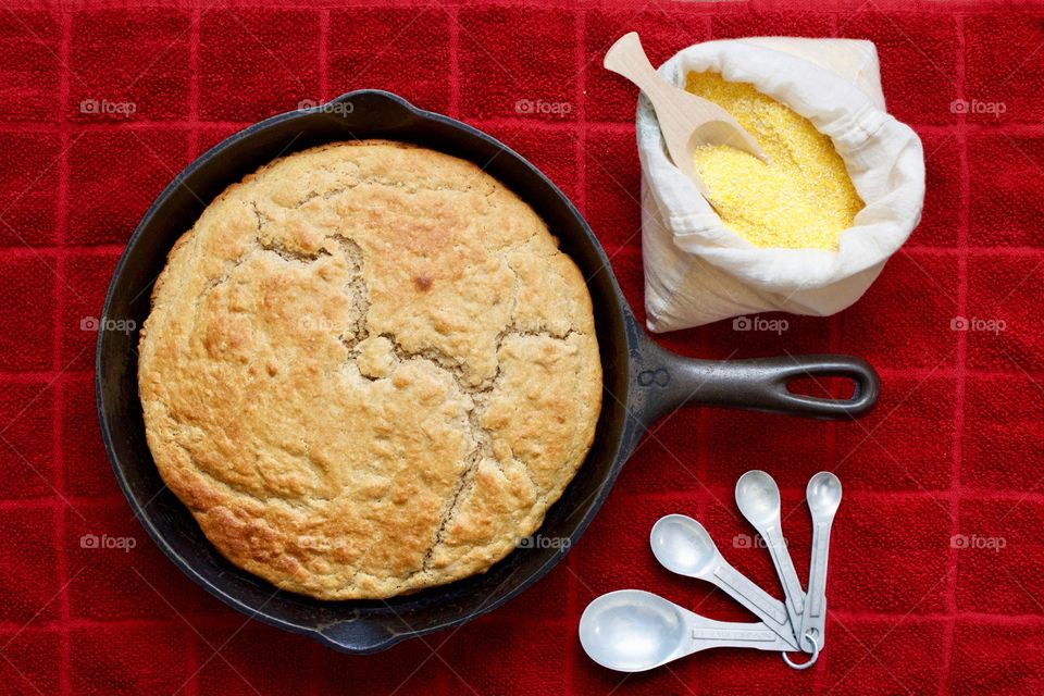 Flat lay of sourdough cornbread in a cast iron skillet, cornmeal and wooden scoop in a flour sack, vintage measuring spoons on a red kitchen towel