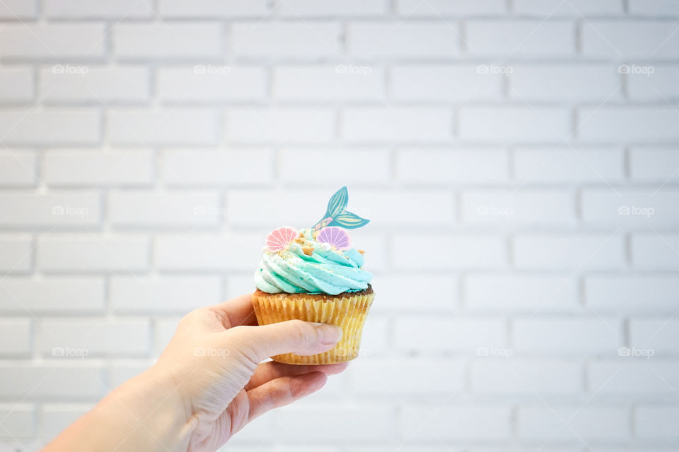 Mermaid cupcake. A lady's hand holding a mermaid muffin against the white brick wall background. fairy tales and fantasy concept. Selective focus on the pink seashell.