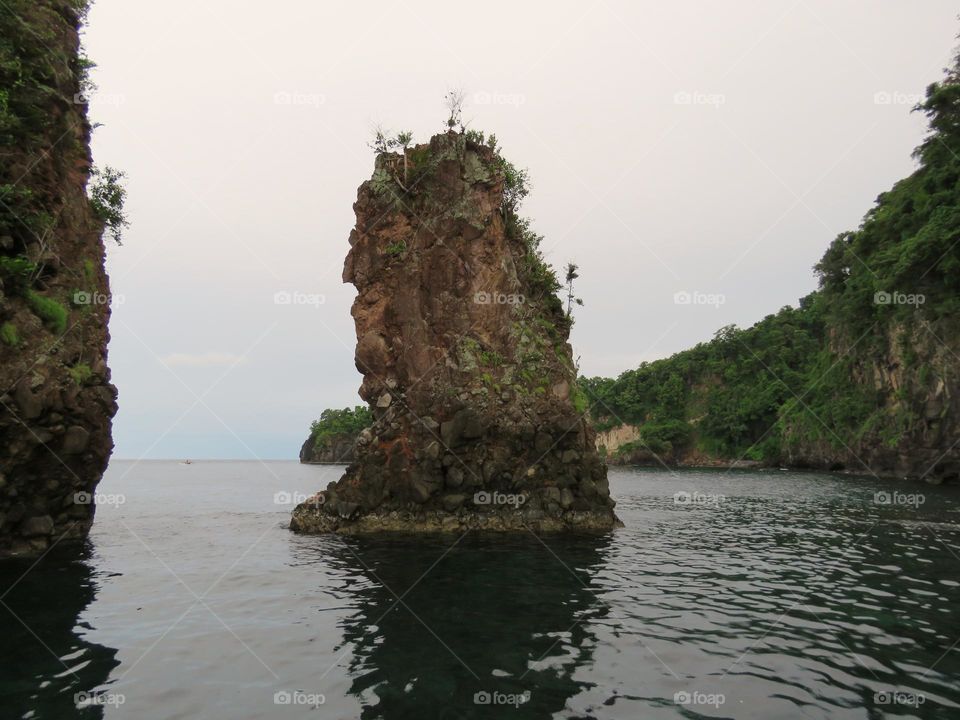 Stone island with green trees on hilly green island background and cloudy sky.