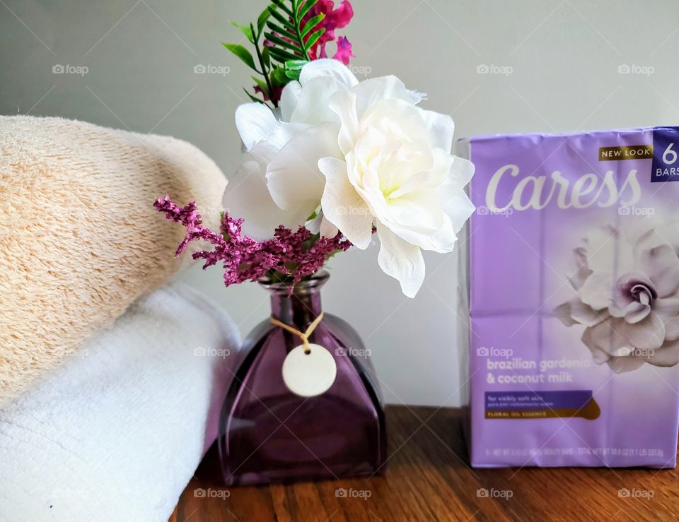 The new look Caress brazilian gardenia & coconut milk six pack bar soap accented by a purple vase and white flowers and with a tan and a white towel stacked on the left.
