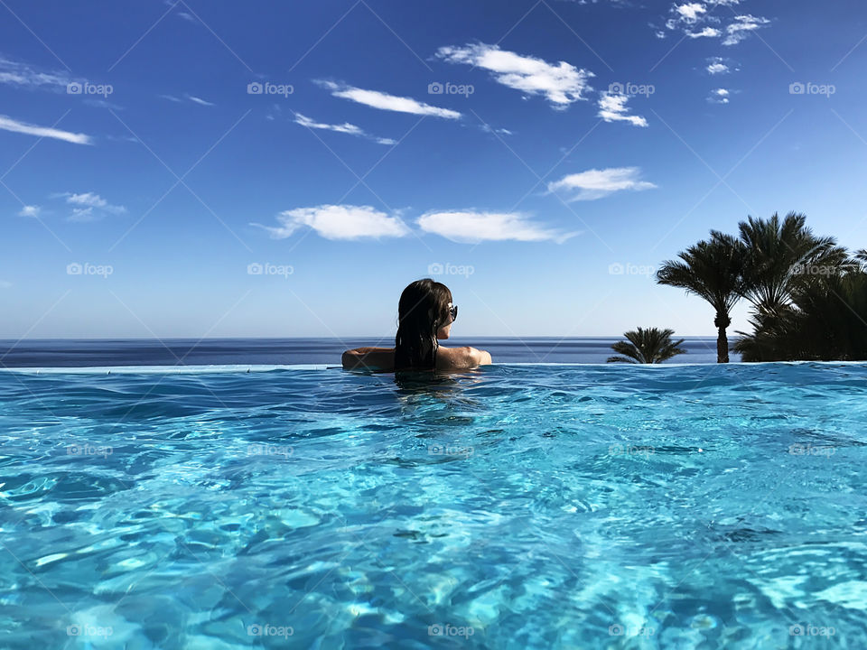 Young woman enjoying the view from the swimming pool to the sea and palm trees on blue sky background 