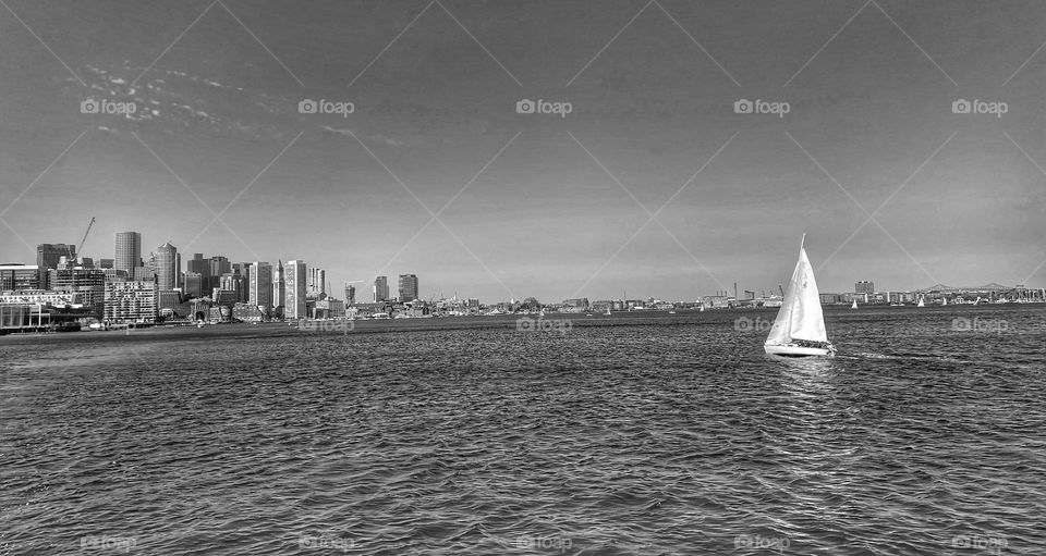 Black and white photo of a line sailboat out on Boston Harbor with the city skyline in the background 