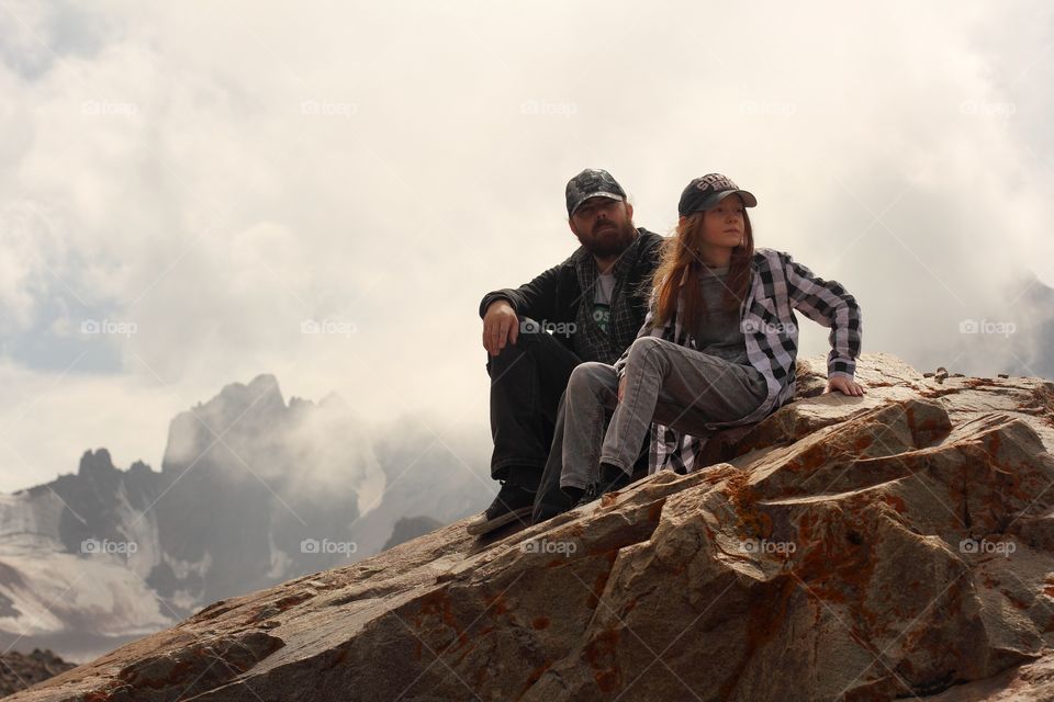 Dad and daughter sit on a rock in the mountains on a glacier in passing clouds