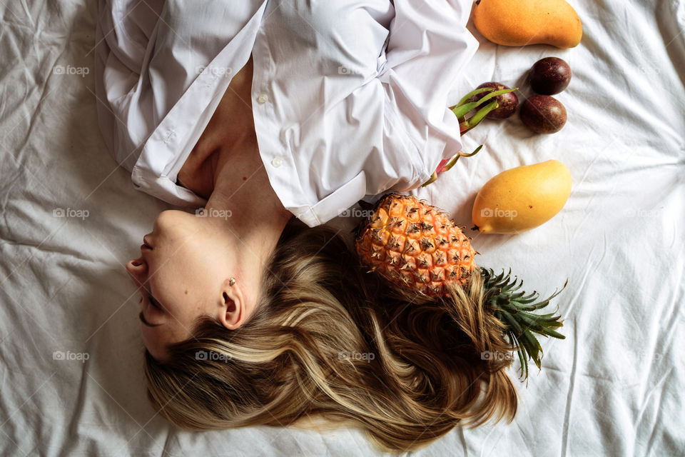 Woman with long blonde hair lying on the bed with fresh exotic fruits