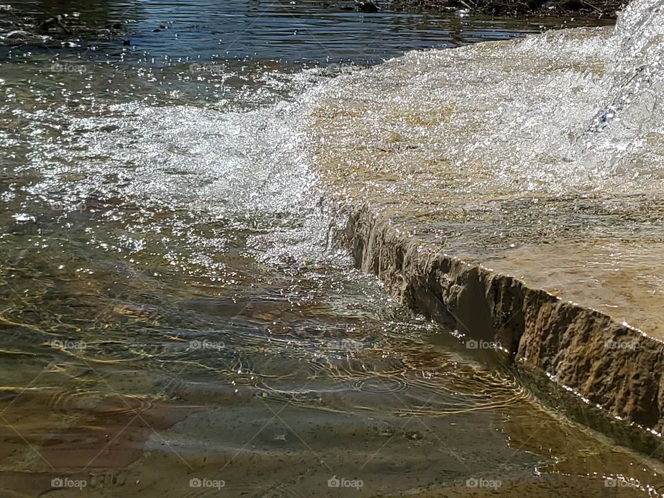 Closeup of a water fountain that fills an urban Spring fed pool with water at a city park. The surface appears to be made out of flagstone and local limestone.