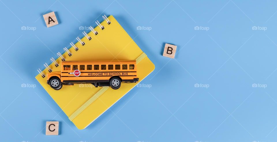 One yellow notebook with a school bus and wooden squares with letters lie on the left on a blue one with copy space on the right, flat lay close-up. Back to school blue and yellow concept.