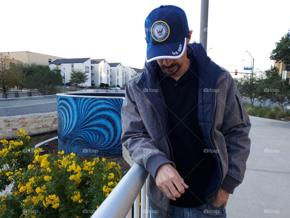 Blue and yellow spotted in the city. A man wearing a blue and yellow Navy baseball cap. He is leaning on a rail next to yellow flowers that are against a blue design tile wall along a city river walkway.