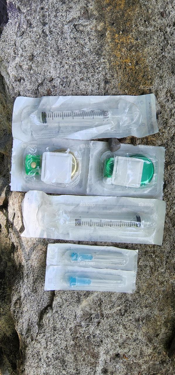 a variety of harm reduction products are found on the forest floor. there are cook plates that are green. there are syringes sealed and there are the complimenting sharps for each syringe. harm reduction for homeless drug addicts in the city