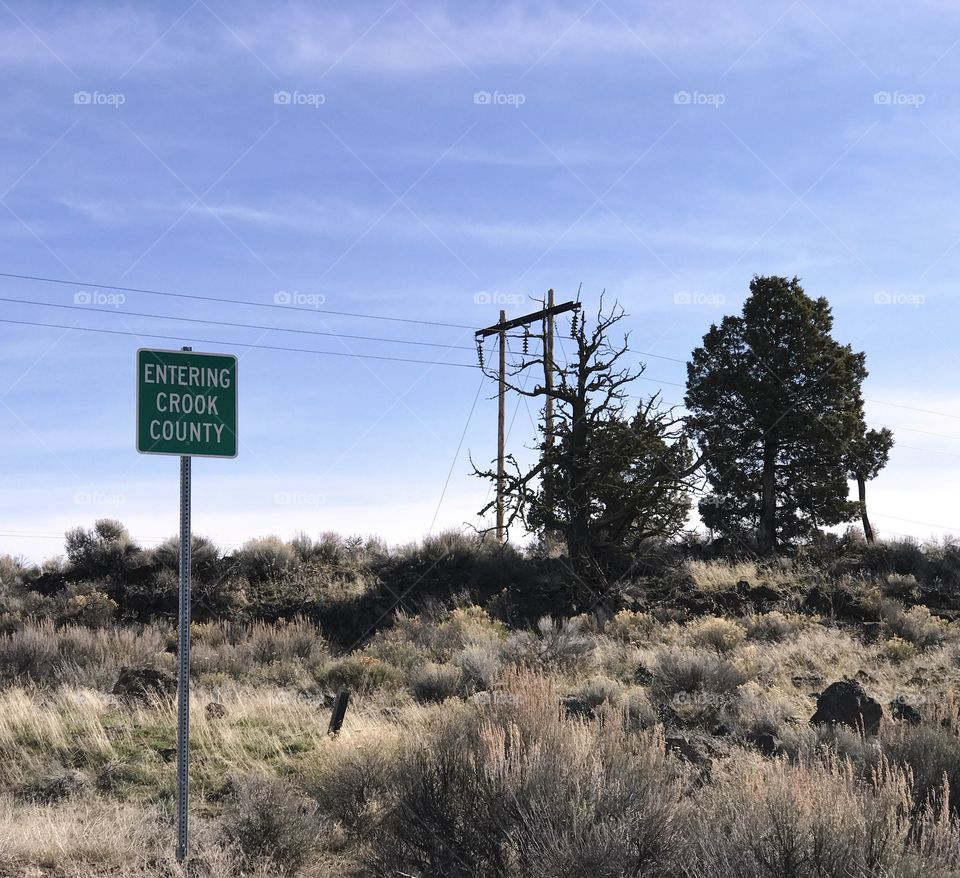 A small sign lets motorists know they are entering rural Crook County in Central Oregon with an area covered in sagebrush, juniper trees, and power poles on a sunny spring day.