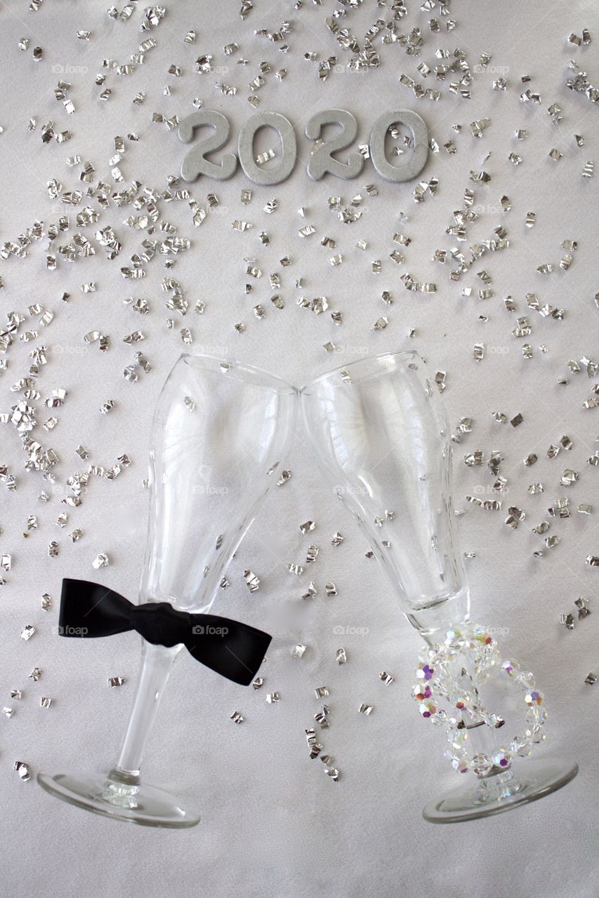 Conceptual overhead view of champagne glasses representing a man and a woman with the year ‘2020’ in silver with silver confetti on a white background - portrait format