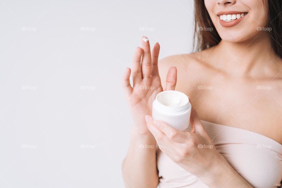 Beauty portrait of happy smiling asian woman with dark long hair put nourishing moisturizer cream on clean fresh skin face and hands on white background
