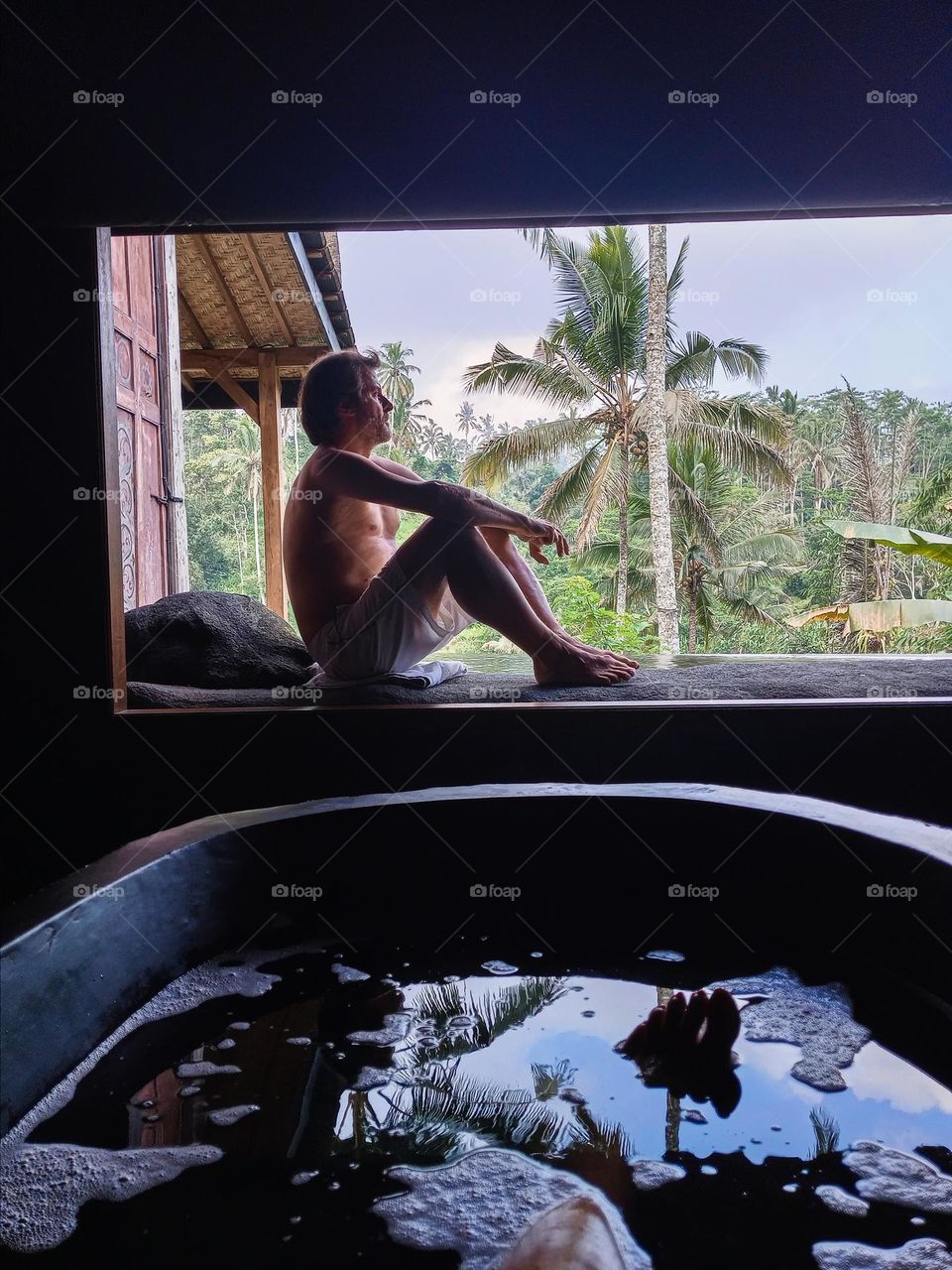 woman in bathtub and man sitting in panoramic window with jungle view
