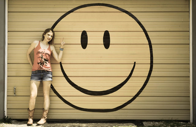 Woman standing in front of a large smiley face mural while giving a peace sign