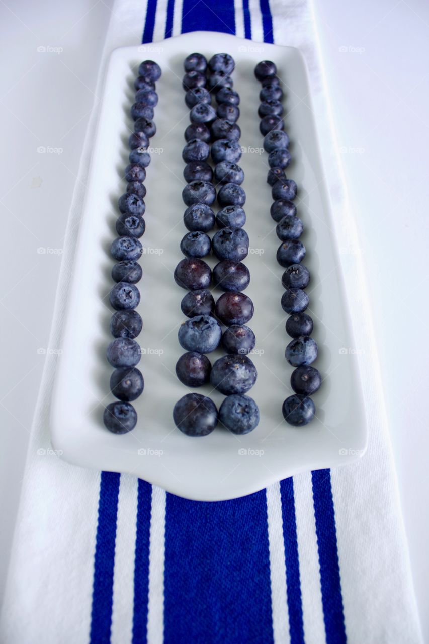 Angled view of blueberries on a rectangular white plate, arranged in lines to emphasize the blue stripes on the white dish towel underneath, all on a white background 