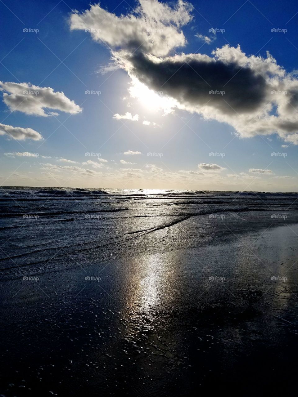 Florida sun setting in the distance over the ocean in Daytona Beach. The shadowed waves lap against the beach. Clouds dot the sky.