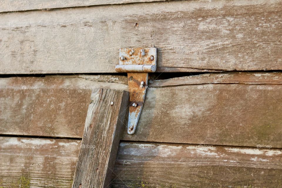 Angled view of a rusted hinge on a weathered wooden ventilation opening in the siding of an antique farm building  