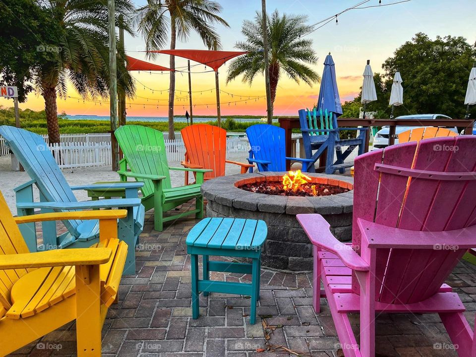 Brightly colored Adirondack chairs around a fire pit at sunset on a beach.