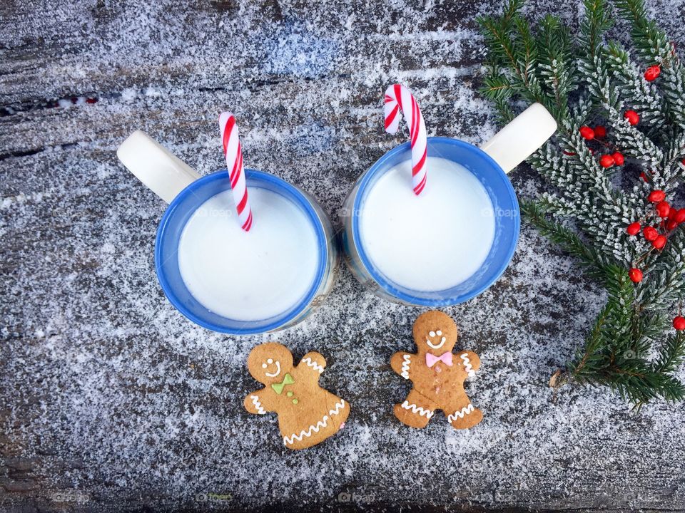 Two mugs of white chocolate and milk with candy canes inside on snowy table with two gingerbread men beside and frozen pine cone tree branches 